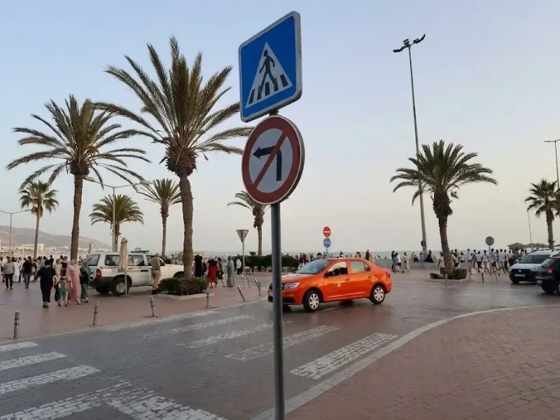 A red petit taxi driving near the Agadir corniche and palm trees during late afternoon, showing the secure tourist area for women researching if Agadir is safe for solo female travellers at night.   